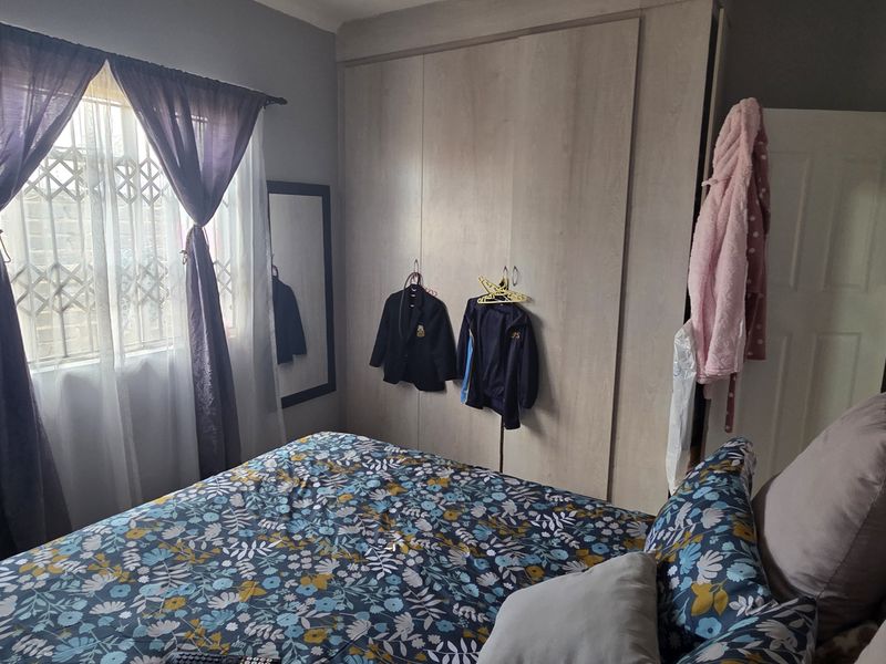 A functional bedroom featuring built-in cupboards and a window with security bars, providing a practical and comfortable living space.