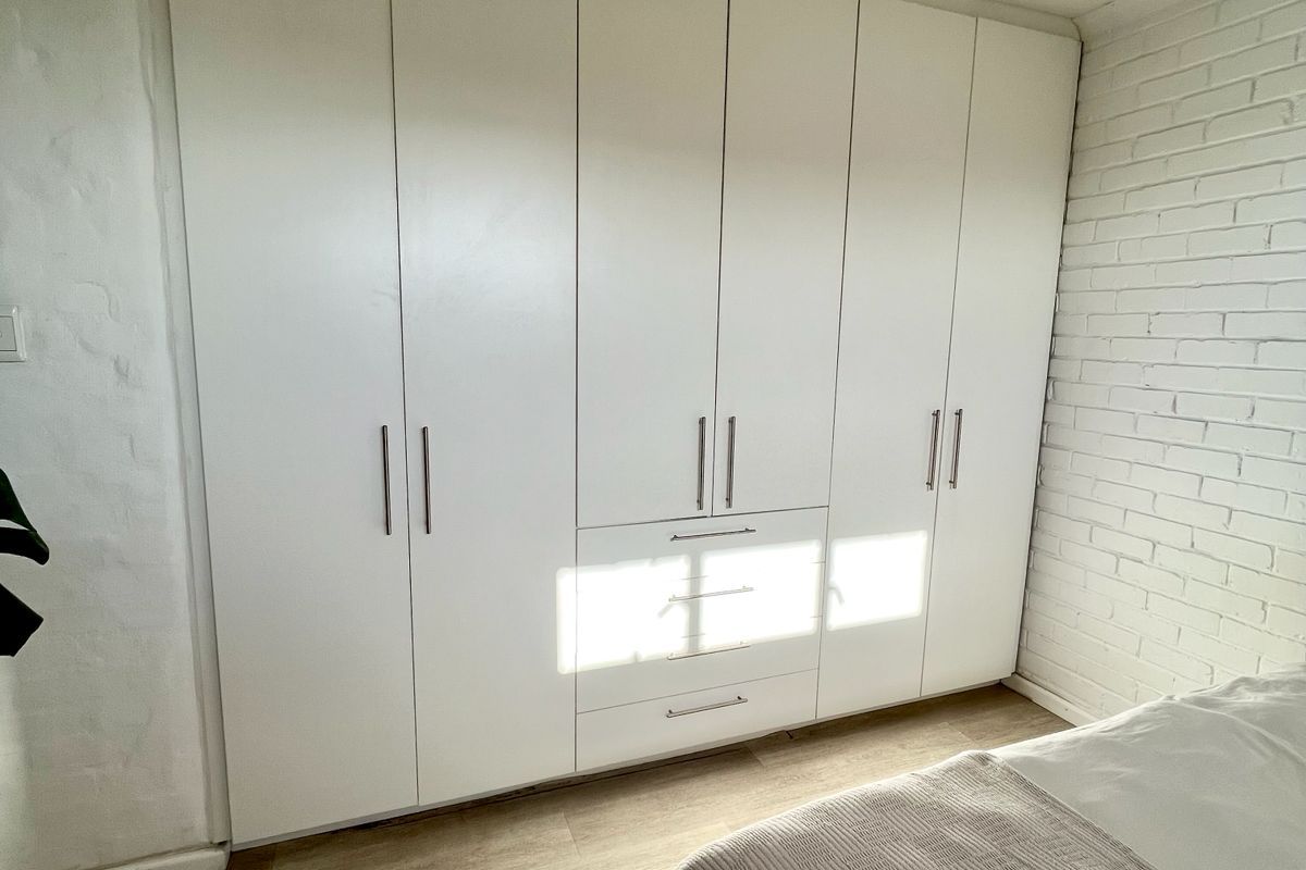 A bright bedroom featuring a large, modern white built-in wardrobe, light laminate flooring, and a textured painted brick feature wall.