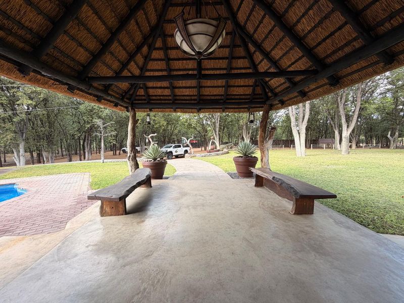 A covered outdoor patio area featuring a traditional thatched roof, exposed timber support posts, and concrete flooring, overlooking a garden and pool area.