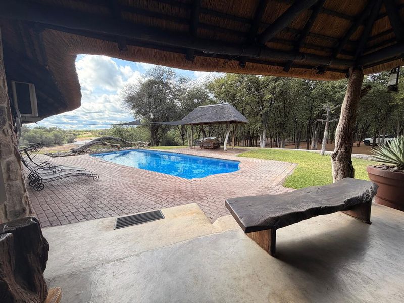 A scenic outdoor pool area featuring a covered thatched patio, brick paving, and a swimming pool overlooking a wooded landscape.