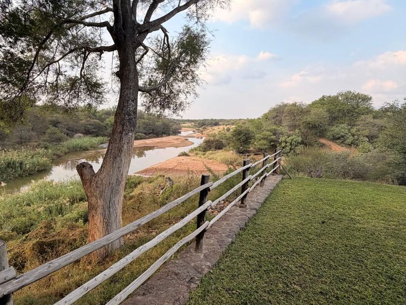 A scenic view overlooking a river landscape, featuring a well-maintained lawn bordered by a stone retaining wall and a timber fence.