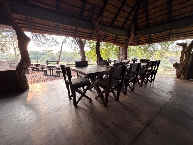 A spacious covered outdoor patio featuring a traditional thatched roof, rustic timber support columns, and a concrete floor overlooking the surrounding landscape.