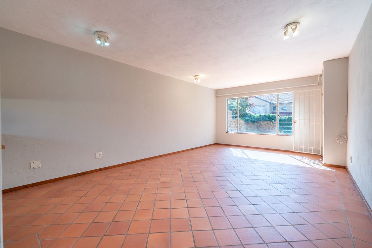 A spacious, empty living area featuring terracotta floor tiles, neutral walls, and a large window with an integrated security gate.