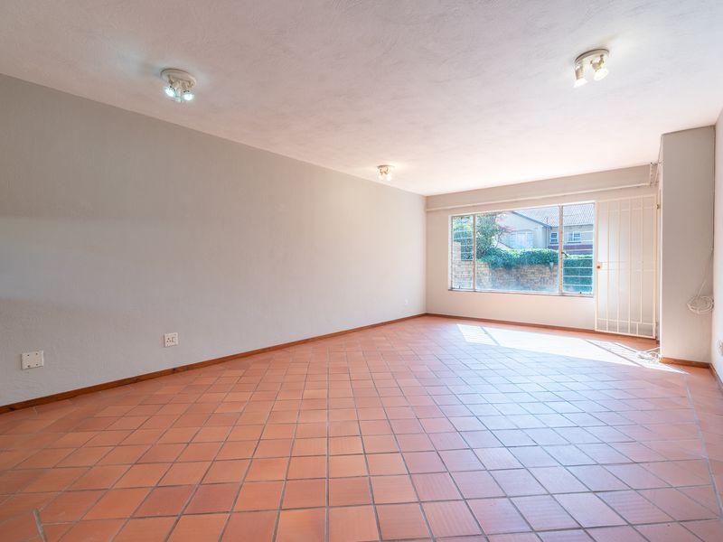 A spacious, empty living area featuring terracotta floor tiles, neutral walls, and a large window with an integrated security gate.