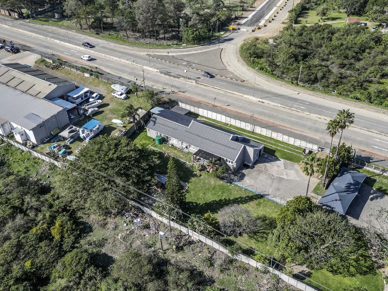 Aerial view of the property showing the main dwelling, driveway, carport, and surrounding garden area adjacent to a main road.