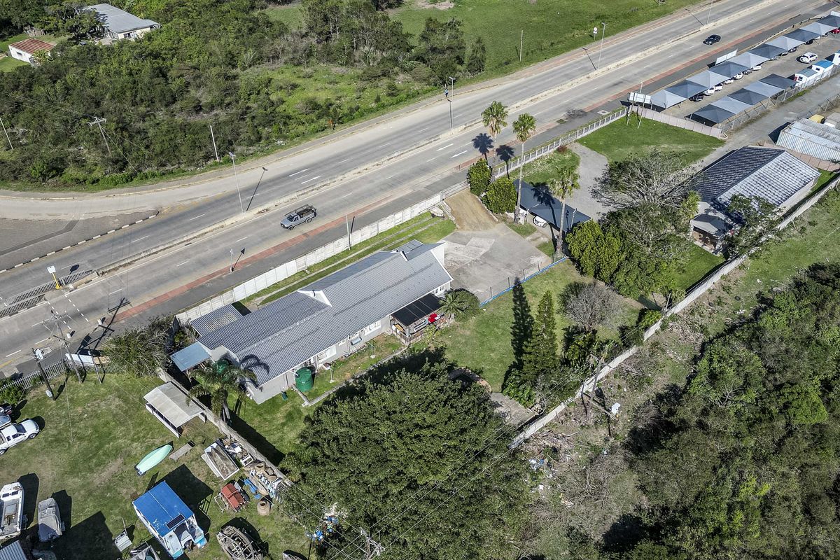 Aerial view of the property showing the main dwelling, paved driveway, perimeter fencing, and surrounding garden area adjacent to the main road.