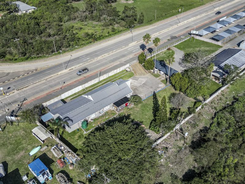 Aerial view of the property showing the main dwelling, paved driveway, perimeter fencing, and surrounding garden area adjacent to the main road.