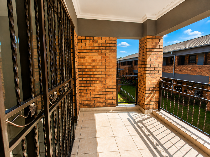 A well-maintained covered balcony featuring tiled flooring, face brick pillars, and decorative wrought iron security features overlooking the apartment complex grounds.