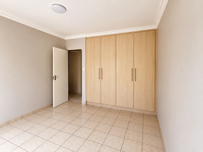 A clean, empty bedroom featuring neutral tiled flooring, light-toned built-in cupboards, and crisp white walls, offering a versatile space ready for personalization.