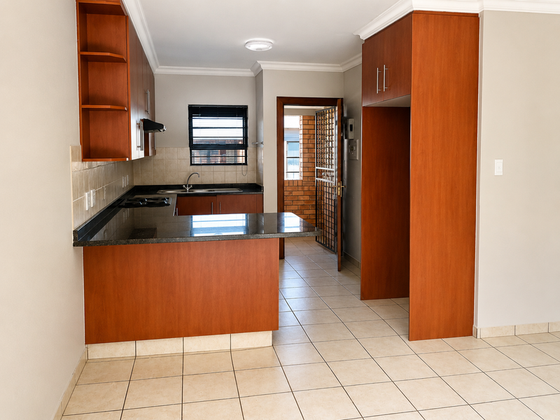 A clean, empty kitchen featuring wood-finish cabinetry, granite countertops, a built-in hob, and tiled flooring, with direct access to an exterior area.