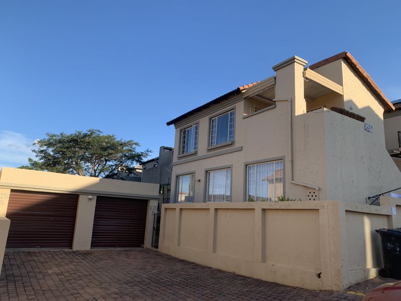 Front exterior view of a double-storey townhouse featuring a double garage with roller shutter doors, paved driveway, and a private balcony.