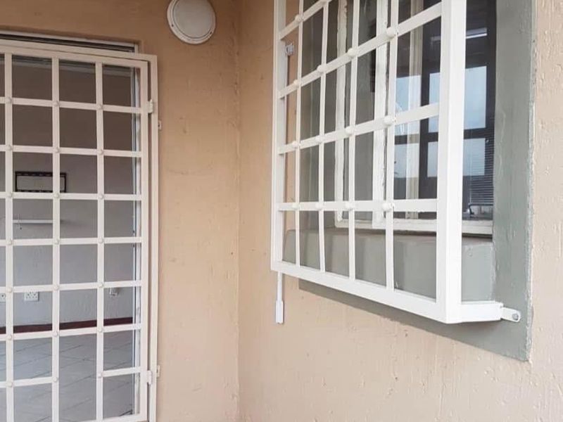 A covered patio area featuring tiled flooring, security gate, and window security bars, showing signs of wear on the exterior wall finish.