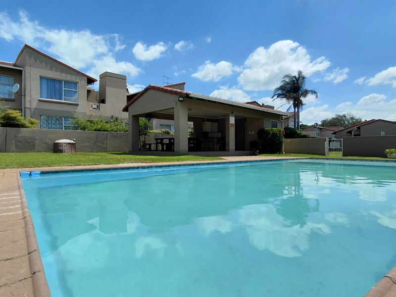A well-maintained outdoor pool area featuring a paved surround, a covered patio structure, and a surrounding lawn with boundary walls.