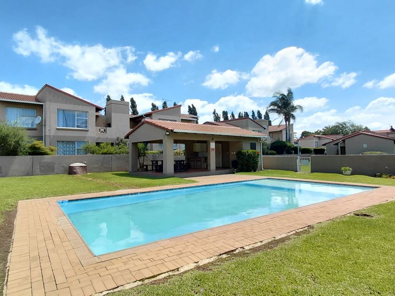 A communal outdoor area featuring a swimming pool with brick coping, a covered patio structure, and well-maintained lawn space within an apartment complex.