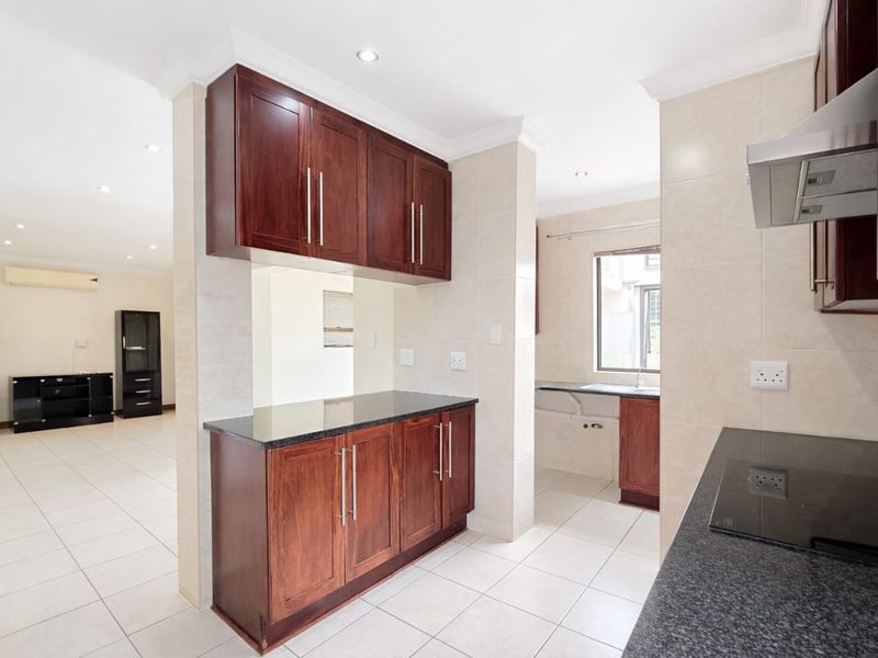 A clean, modern kitchen featuring dark wood cabinetry, granite countertops, integrated appliances, and neutral tiled flooring, flowing into an open-plan living area.