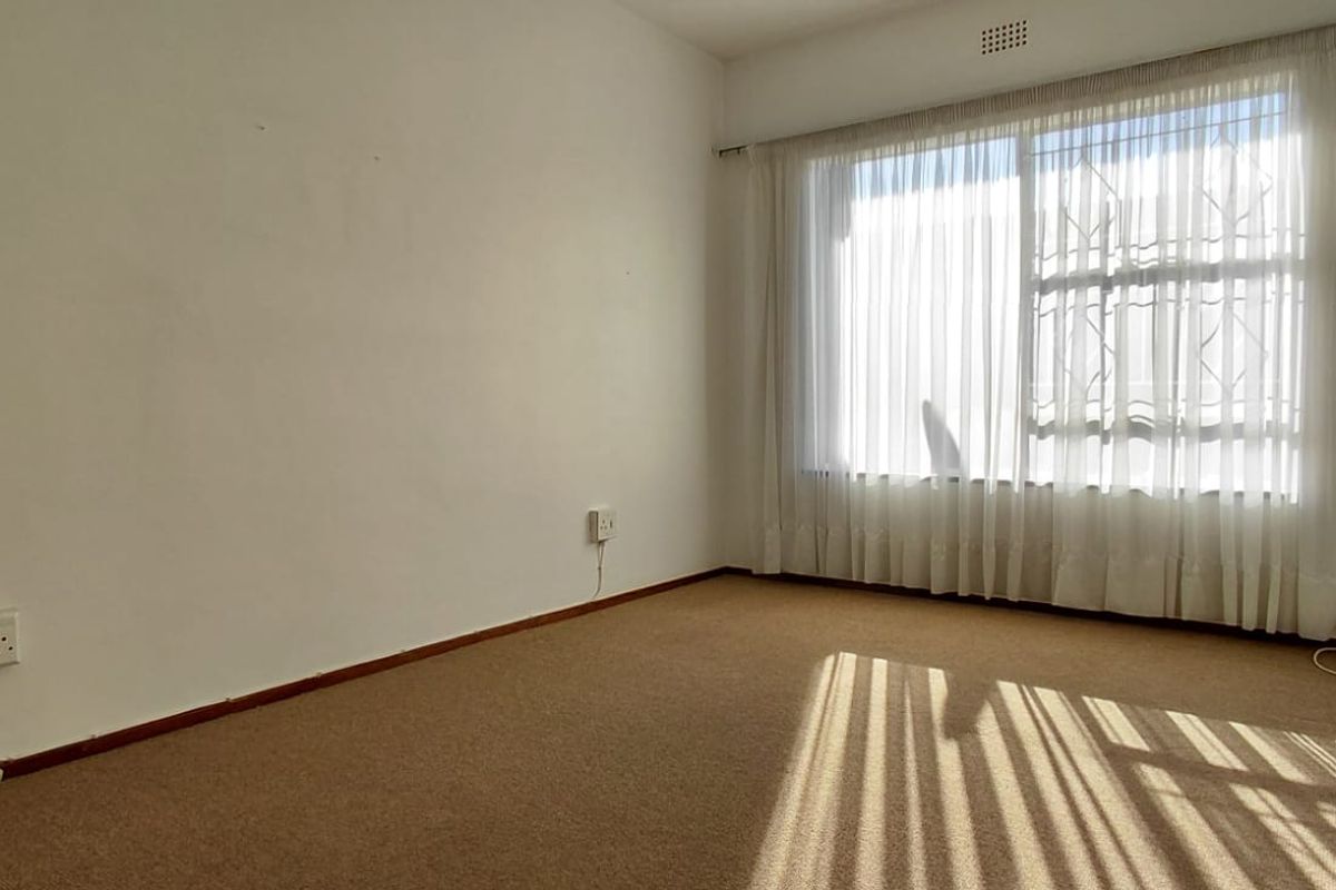 An empty bedroom featuring carpet flooring, a large window with security bars, and neutral walls, offering a blank canvas for renovation.