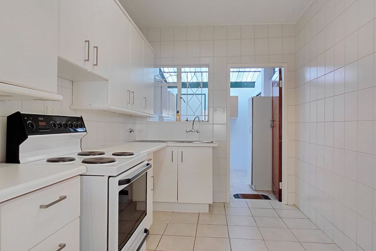 A functional kitchen featuring white built-in cabinetry, a freestanding electric stove, tiled splashbacks, and tiled flooring, with an adjacent utility area.