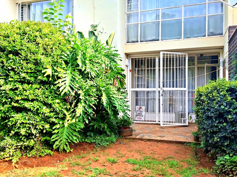 Rear view of the townhouse featuring a paved patio area, security gate access, and large windows, set against a garden space requiring maintenance.