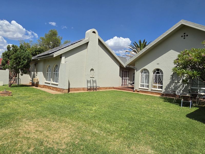 A well-maintained rear garden featuring a spacious lawn, perimeter wall, and the rear facade of the house with arched windows and solar panels.