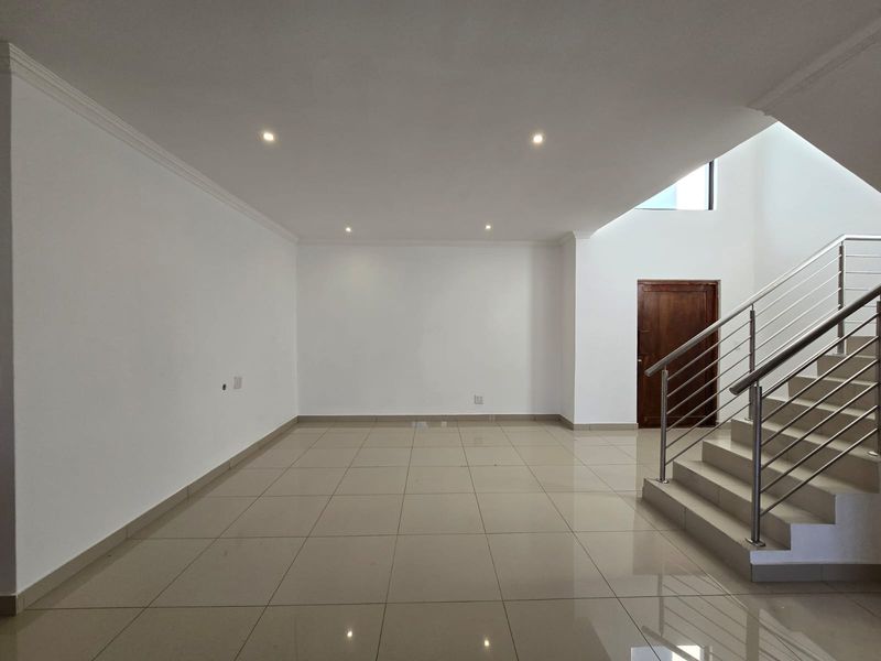 A spacious, open-plan living area featuring neutral tiled flooring, recessed lighting, and a modern staircase with a stainless steel balustrade.