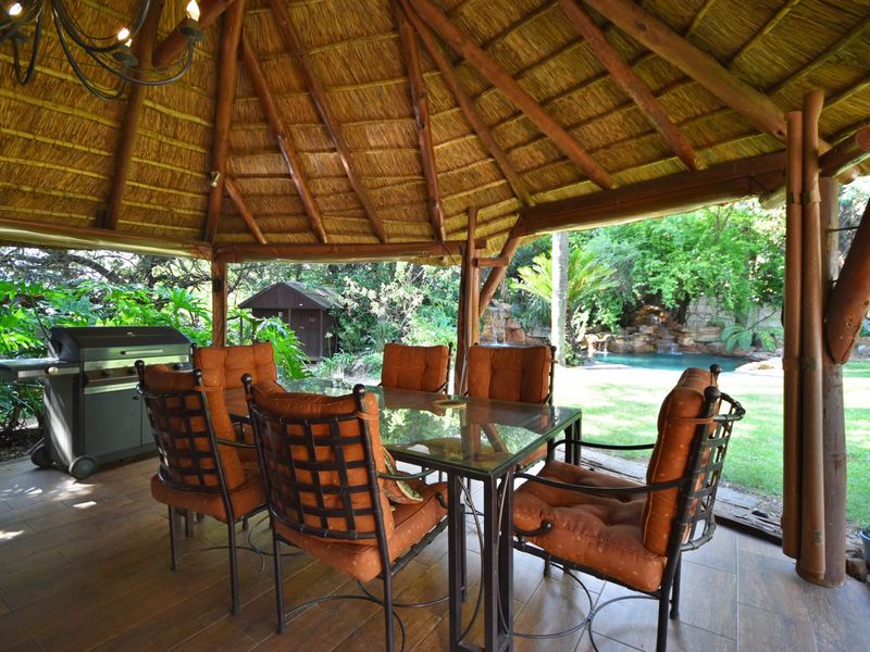 A spacious outdoor thatched-roof patio area featuring tiled flooring and timber support poles, overlooking a lush garden and swimming pool.