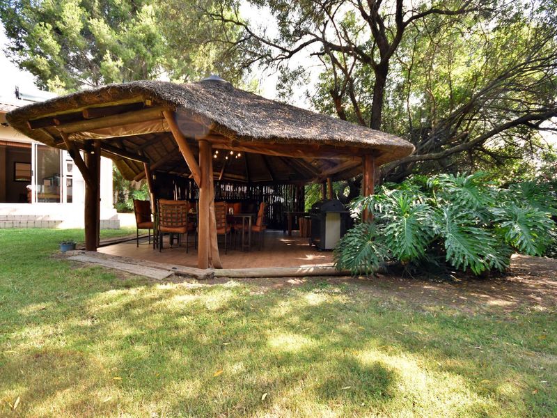 A covered outdoor patio area featuring a traditional thatched roof, timber support structure, and tiled flooring, set within a landscaped garden.