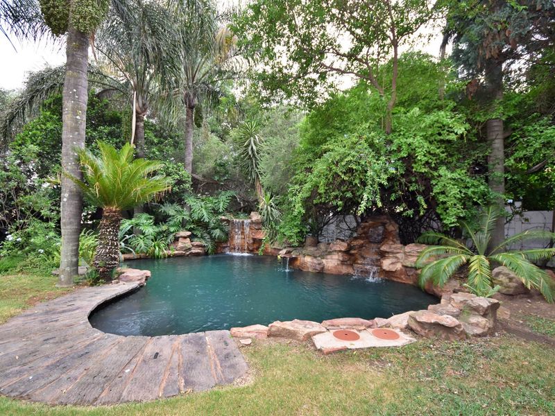 A naturalistic rock-feature swimming pool set within a lush, mature garden, featuring integrated waterfalls and timber decking.