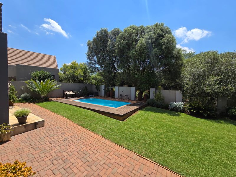 A well-maintained backyard featuring a swimming pool with timber decking, brick paved patio area, and established garden landscaping.