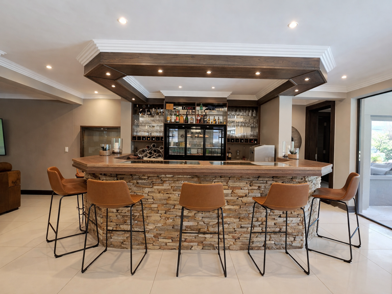 A well-appointed home bar featuring a stone-clad counter, custom wooden bulkhead with integrated lighting, and built-in beverage refrigeration.