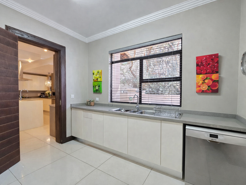 A well-maintained scullery featuring modern built-in cabinetry, stone countertops, a stainless steel sink, and tiled flooring, with a view into the adjacent kitchen.