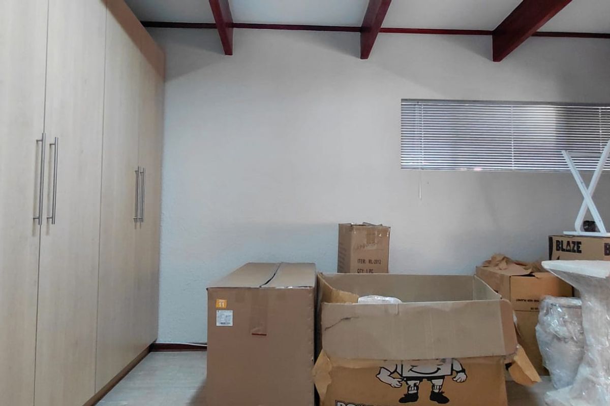 A bedroom featuring light wood-finish built-in cupboards, exposed ceiling beams, and venetian blinds, currently used for storage.