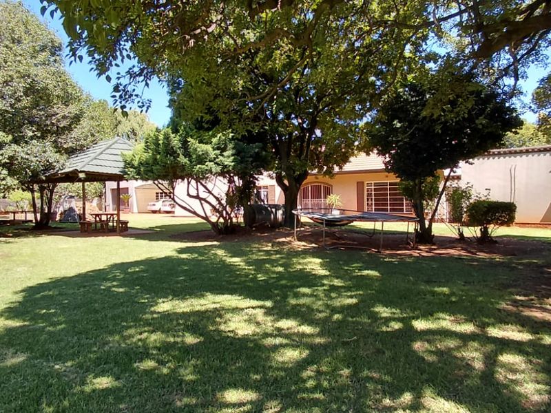 A spacious, well-established garden featuring mature trees, a manicured lawn, a boundary wall, and a charming thatched gazebo for outdoor relaxation.