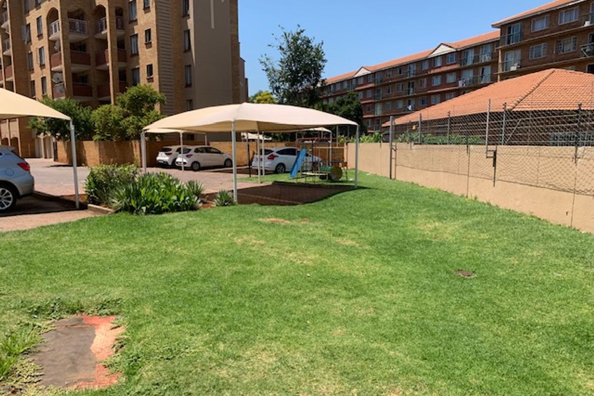 Communal garden area featuring a shade sail structure, perimeter walling, and paved parking access within the apartment complex grounds.