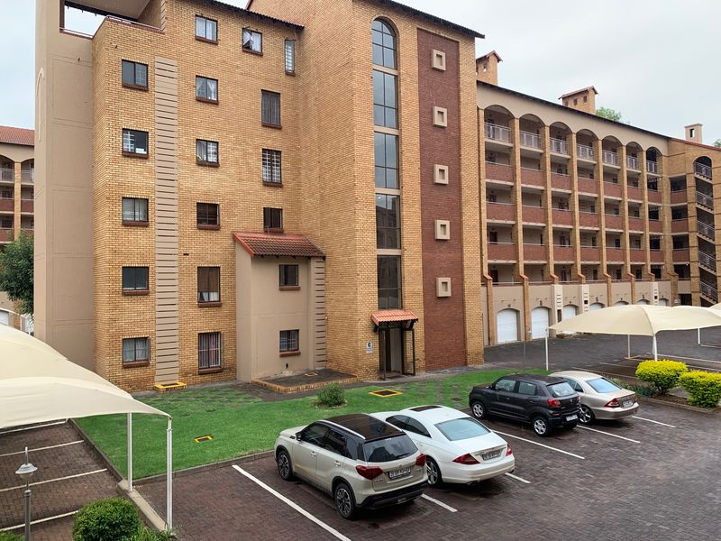 Exterior view of a multi-story apartment complex featuring face brick architecture, paved parking bays with shade cloth carports, and landscaped garden areas.