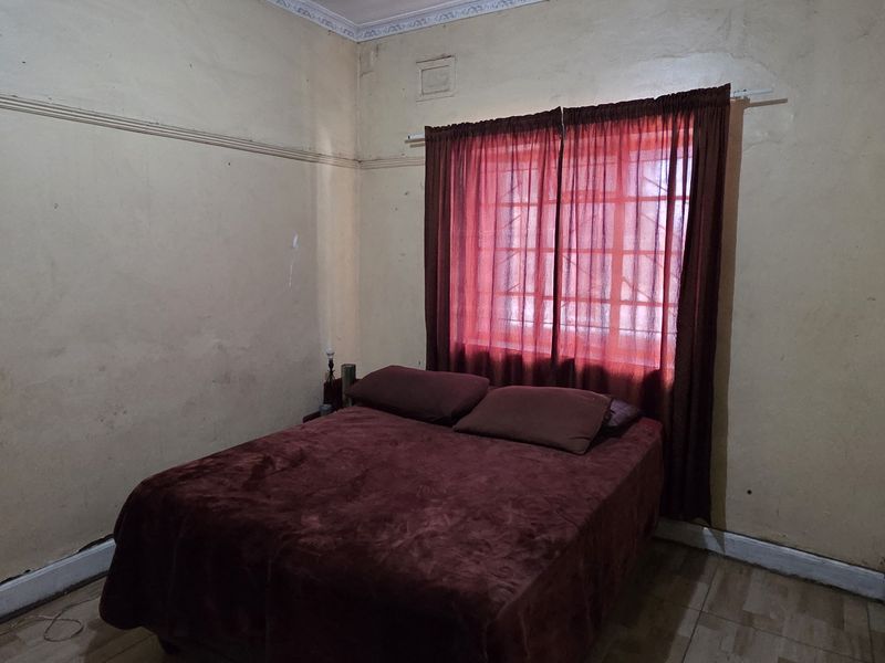 A bedroom featuring tiled flooring, a built-in wardrobe, and a window with security bars. The room requires cosmetic updates to the wall finishes.