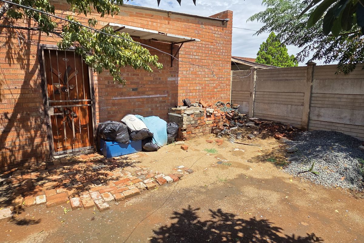 Rear exterior area featuring a brick facade, security gate, and precast boundary wall, currently requiring significant clearing and landscaping to improve curb appeal.