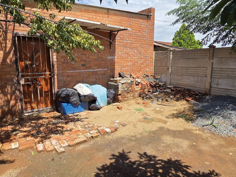 Rear exterior area featuring a brick facade, security gate, and precast boundary wall, currently requiring significant clearing and landscaping to improve curb appeal.