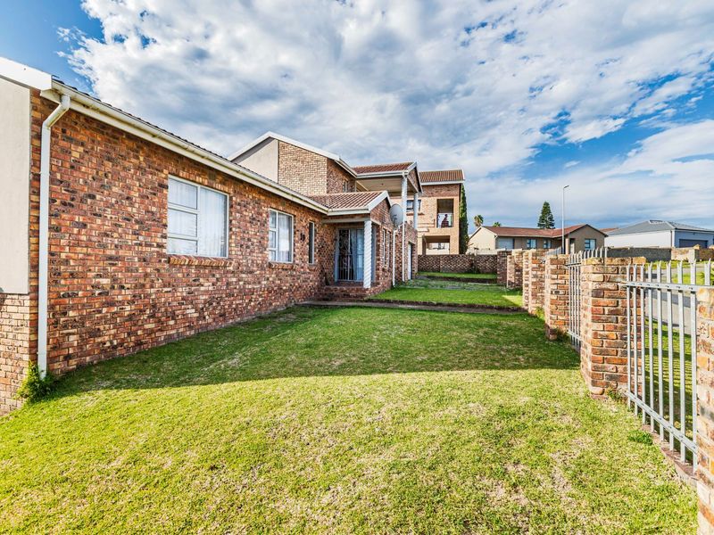 A view of the front exterior of a face brick house featuring a well-maintained lawn, brick boundary walls, and metal security fencing.