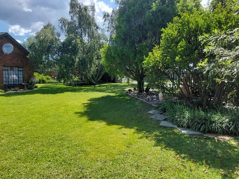 A spacious, well-maintained garden area featuring a lush green lawn, mature trees, and a brick-faced exterior wall of the house.
