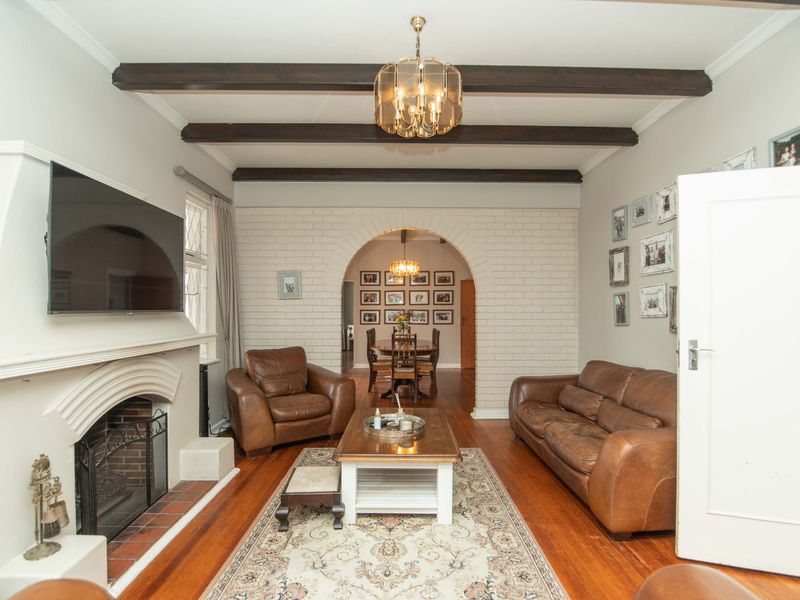 Cozy living room featuring exposed timber ceiling beams, a classic brick fireplace, and hardwood flooring, leading through an arched doorway to the dining area.