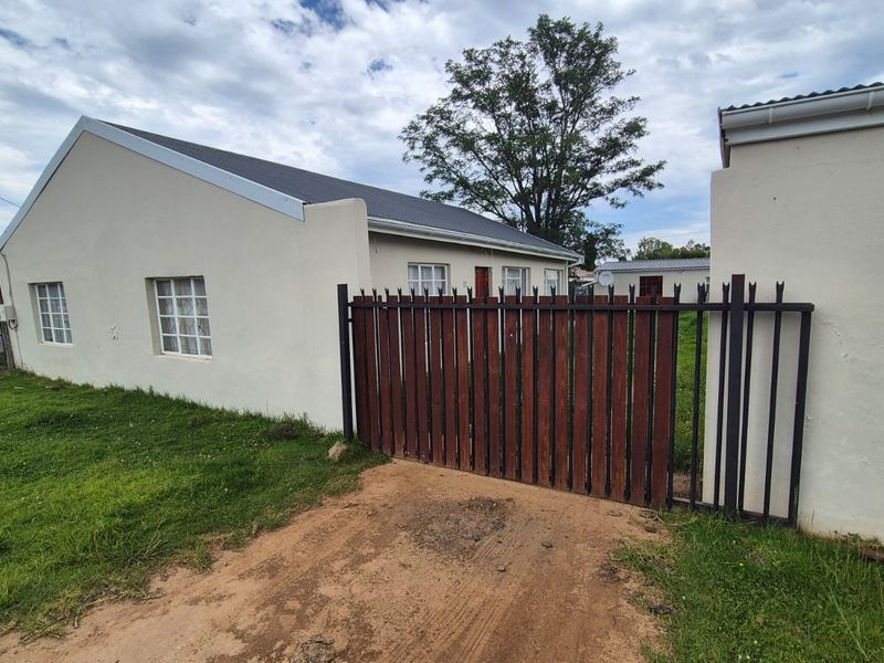 Front exterior view of a residential house featuring a metal security gate, pitched roof, and an unpaved driveway entrance.