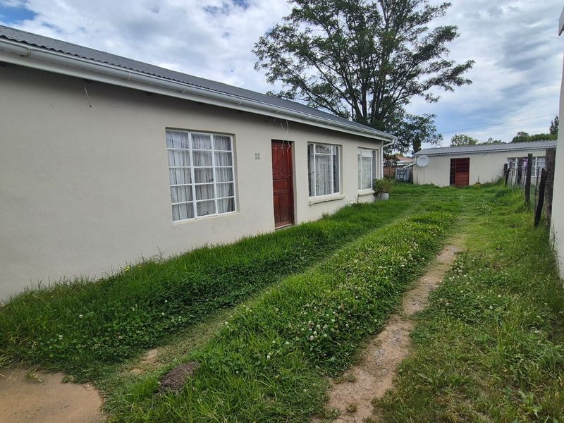 Rear view of the property showing the exterior facade, corrugated metal roof, and an expansive, unmaintained grass area.