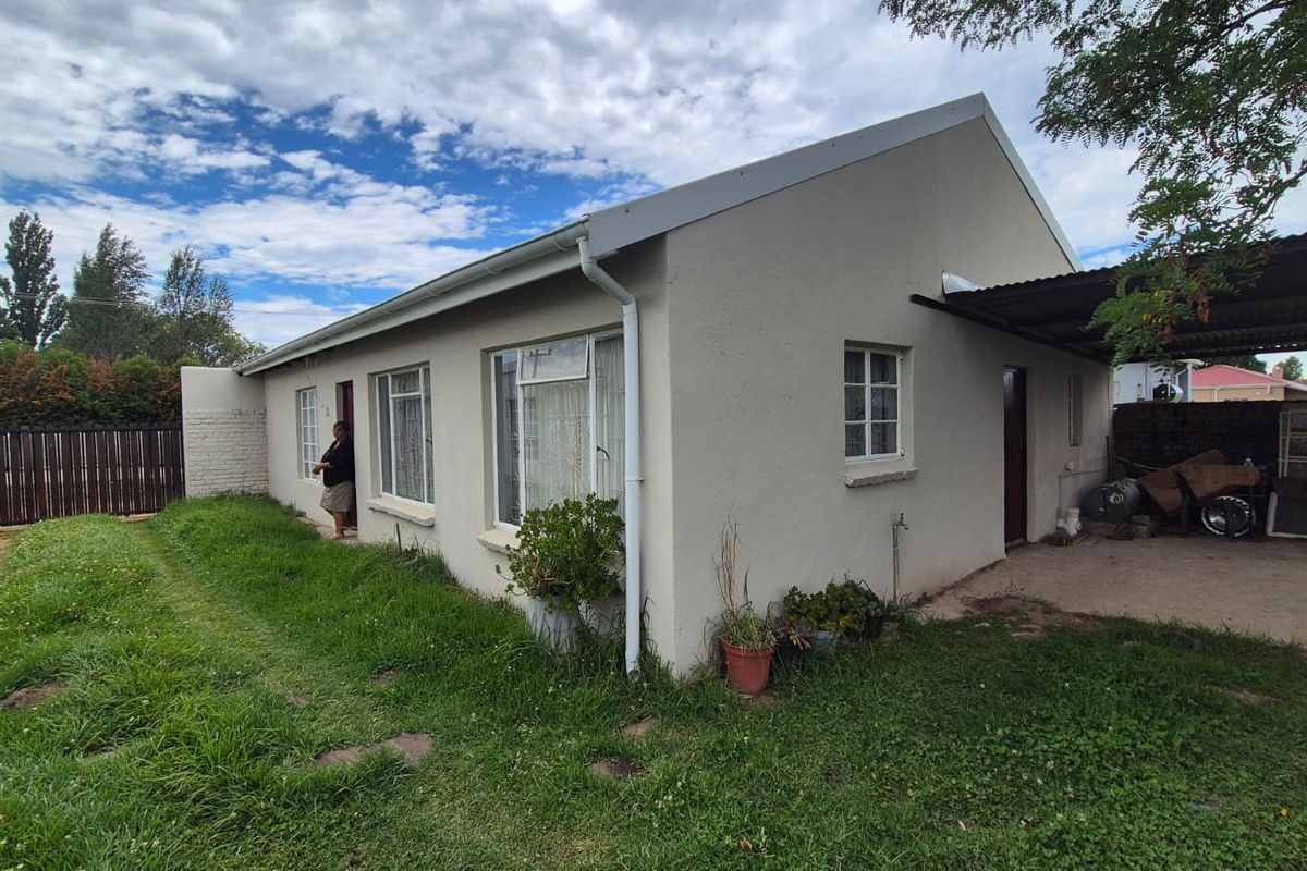 Rear view of the property featuring a simple facade, attached carport, and surrounding garden area with established lawn.