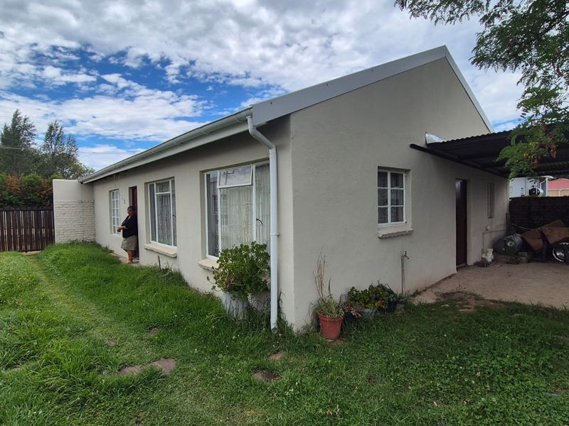Rear view of the property featuring a simple facade, attached carport, and surrounding garden area with established lawn.