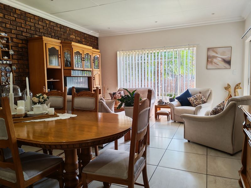 Spacious open-plan living and dining area featuring a rustic exposed brick wall, neutral tiled flooring, and large windows with vertical blinds.