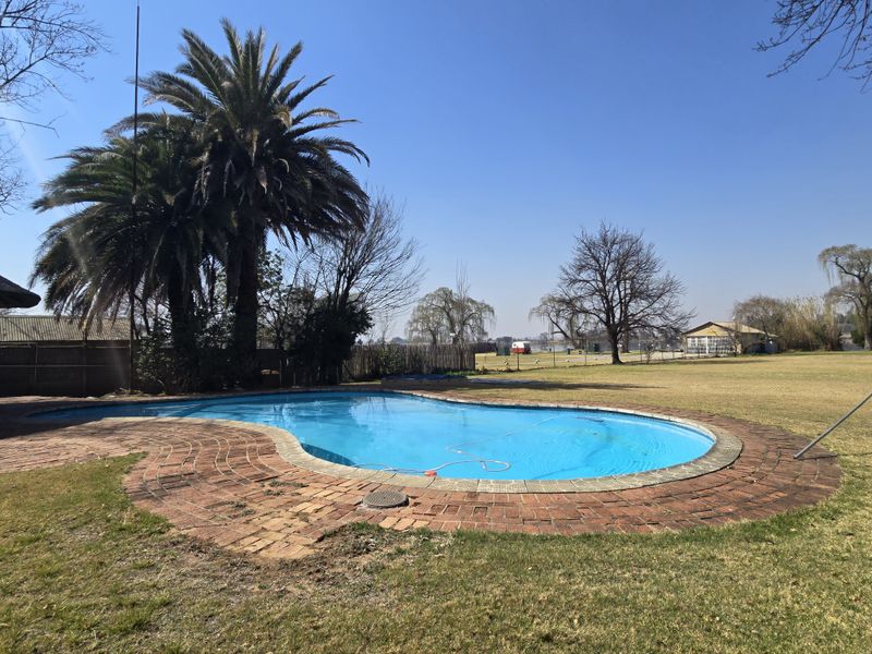 A spacious outdoor pool area featuring a curved swimming pool surrounded by brick paving, set within a large, open garden landscape.