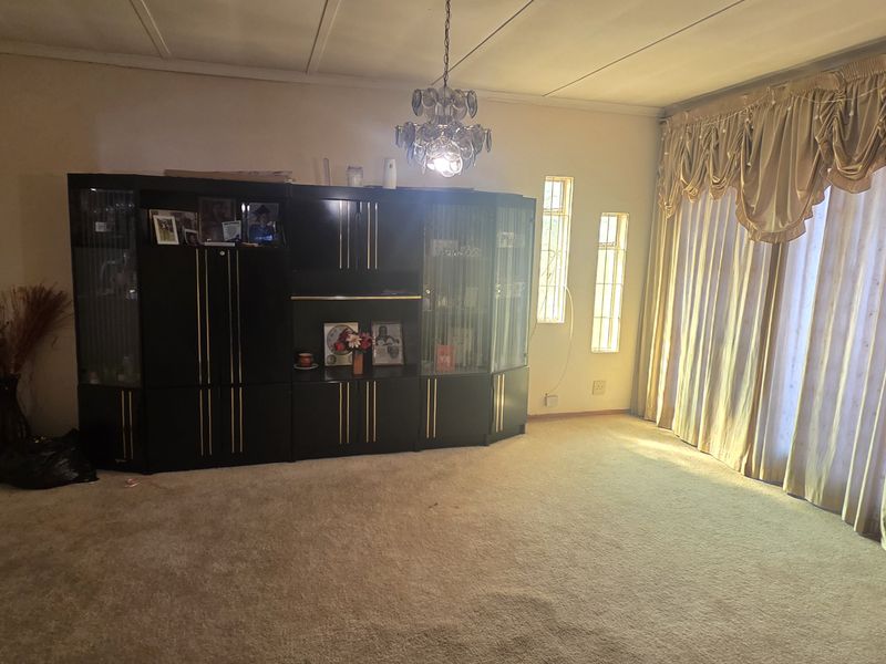 A spacious living room featuring carpeted flooring, a central chandelier, and large windows with draped curtains, offering a comfortable and traditional living space.