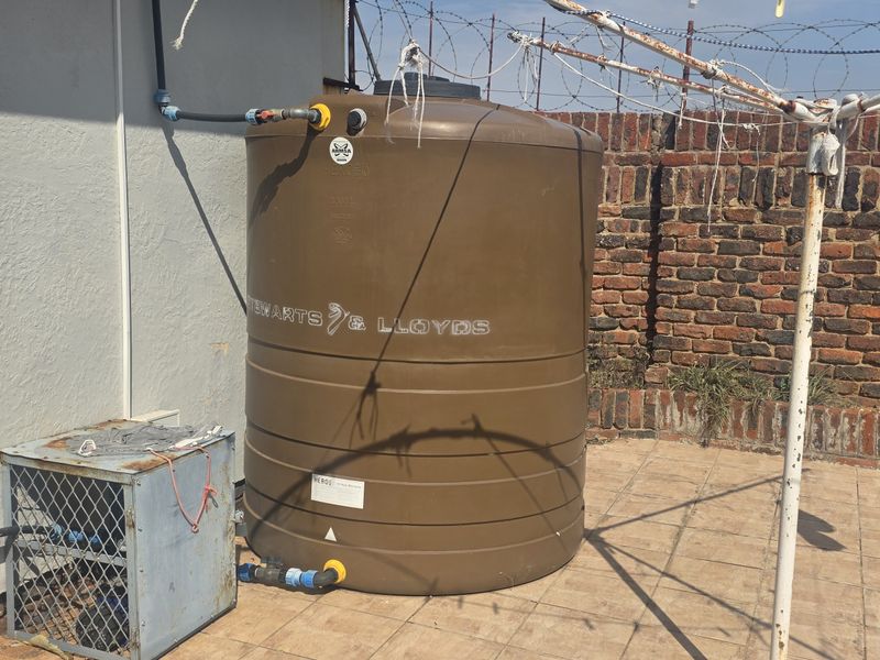 Outdoor utility area featuring a large water storage tank, plumbing infrastructure, and a brick boundary wall on a tiled patio surface.