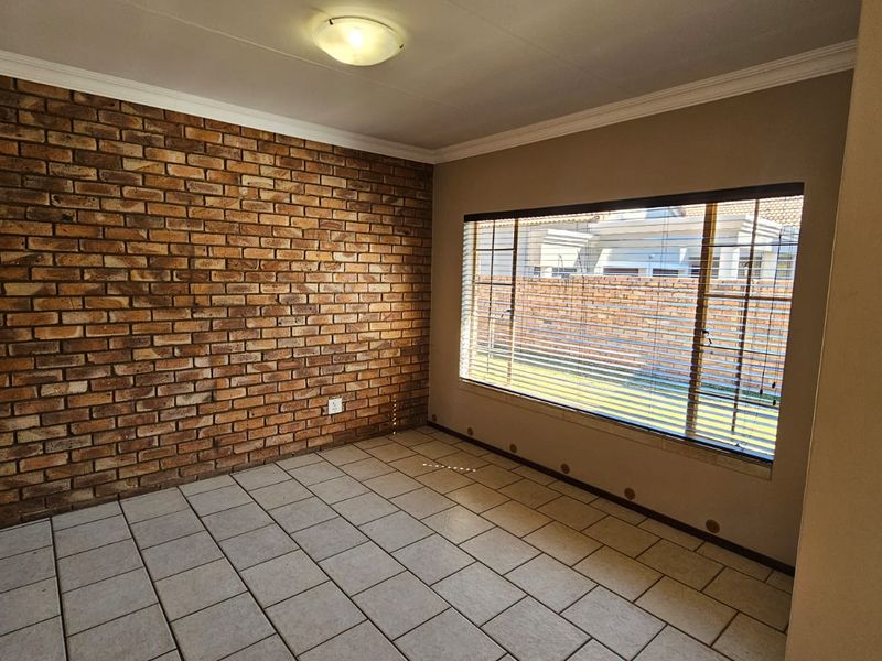 An empty living room featuring a prominent exposed brick wall, ceramic tile flooring, and a large window providing natural light.