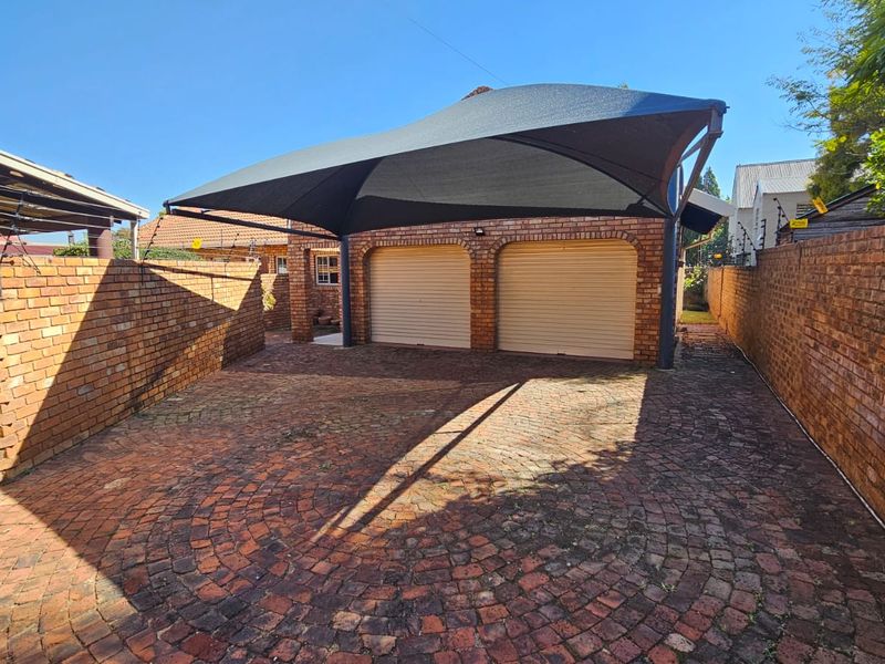 A double garage with roll-up doors, featuring a shade sail carport and a brick-paved driveway enclosed by perimeter walls with electric fencing.